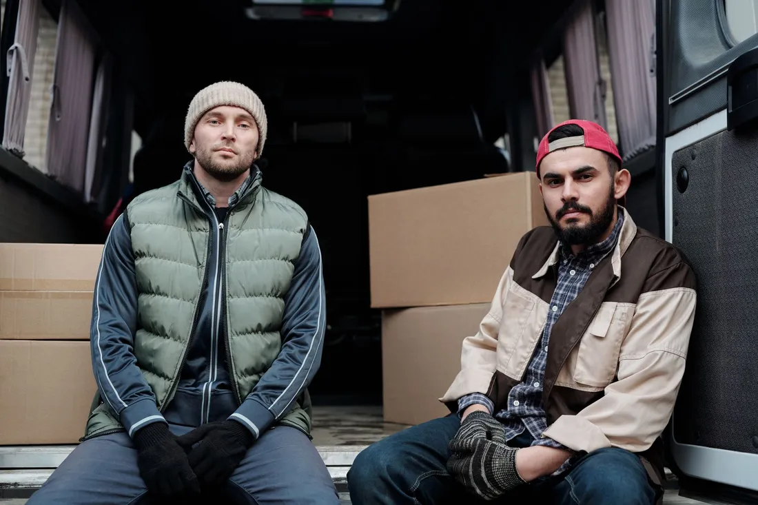 Zwei Umzugshelfer mit Arbeitshandschuhen sitzen in der offenen Ladefläche ihres Transporters neben gestapelten Kartons.
