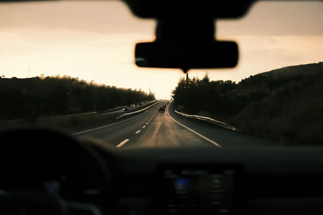 Blick durch die Windschutzscheibe auf eine leere Autobahn in der Abenddämmerung, am Horizont flache Hügel.