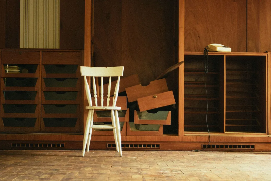 Alter Einbauschrank mit herausgezogenen Schubladen, davor ein heller Holzstuhl auf abgenutztem Parkett.