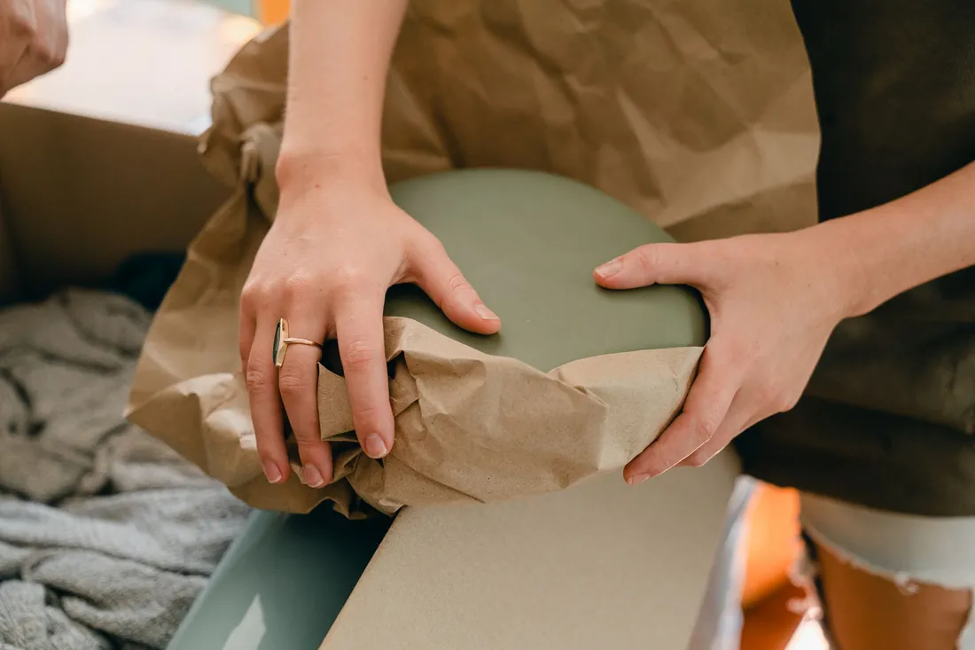 Zwei Hände schlagen ein zerbrechliches Teil in Packpapier ein, daneben steht ein offener Karton.
