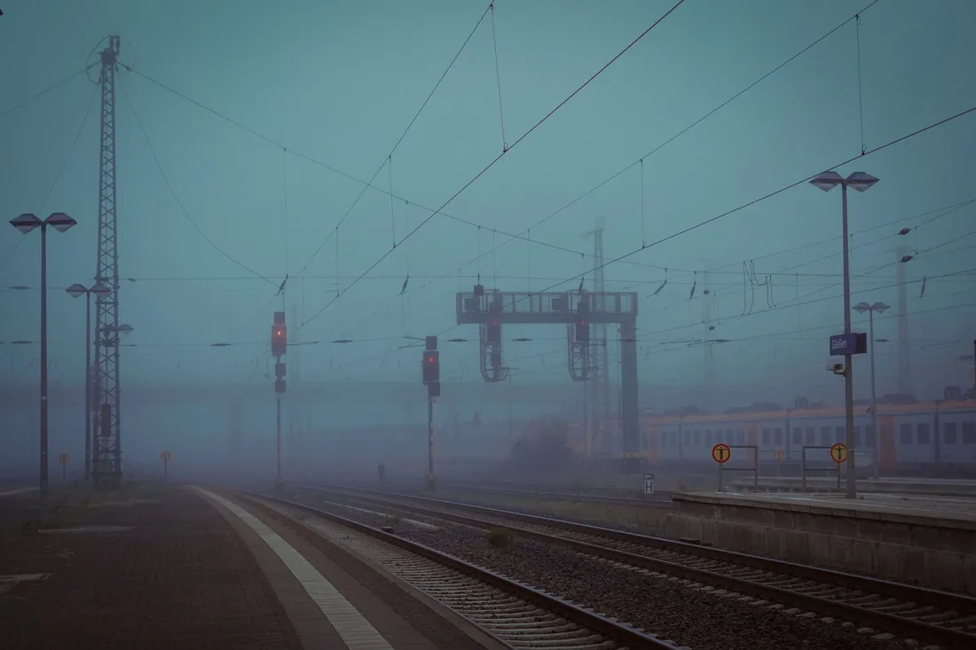 Nebliger Bahnsteig im Morgengrauen mit Gleisen, Oberleitungen und einem wartenden Regionalzug.