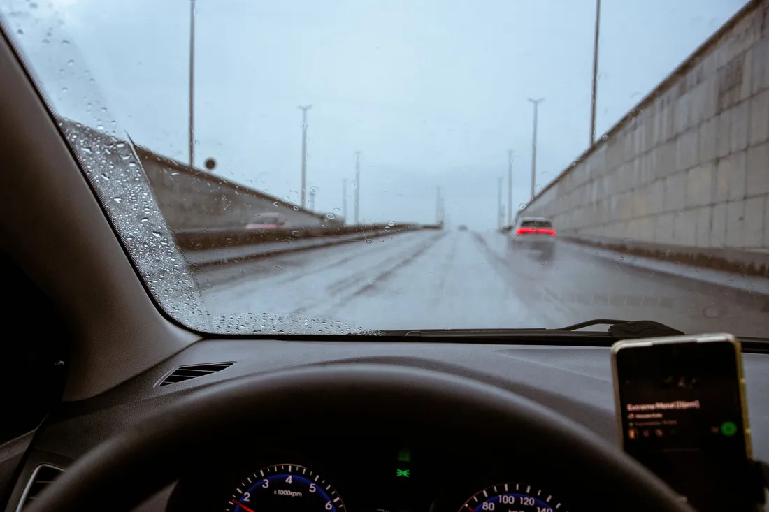 Blick durch die regennasse Windschutzscheibe auf eine Autobahn mit Schallschutzwand und Rücklichtern.