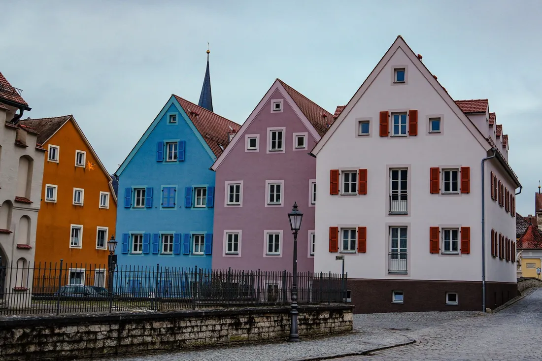 Zeile bunter Giebelhäuser mit Fensterläden an einer gepflasterten Altstadtstraße, dahinter ein Kirchturm.