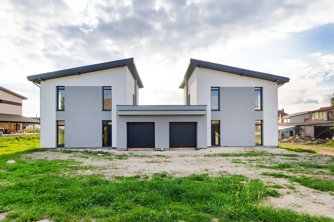 Zwei neu gebaute Doppelhaushälften mit hellem Putz, dunklen Fenstern und Garagentoren am Ortsrand.