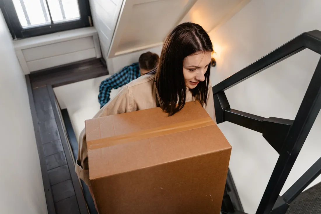 Zwei Personen tragen große Umzugskartons über eine Treppe nach oben, aufgenommen von oben.