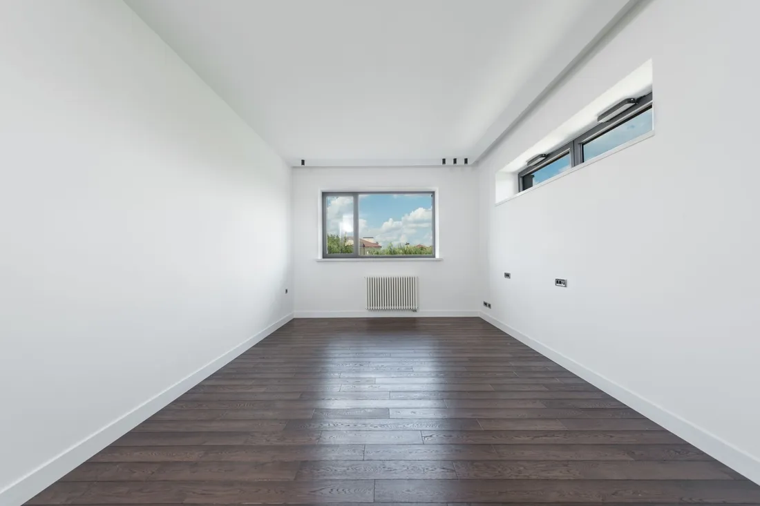 Völlig leerer Wohnraum mit dunklem Parkett, weißen Wänden, Fenster und Heizkörper darunter.